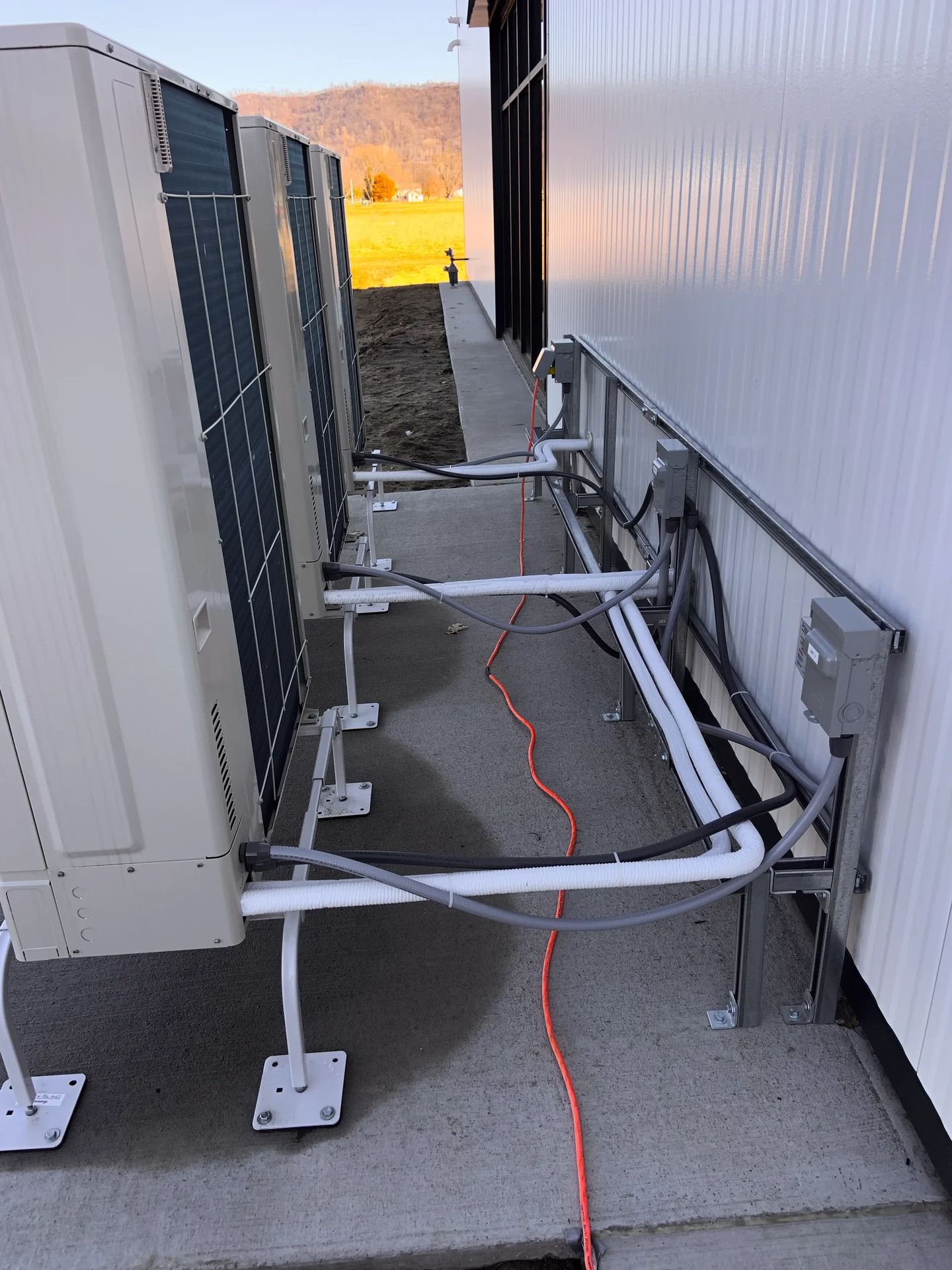 A row of heat pump outdoor units mounted on a wall rack with line sets and disconnects